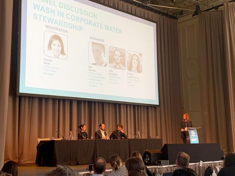 There is a stage with a large monitor displaying a slide deck that says Panel Discussion - WASH in Corporate Water Stewardship. Cheryl Hicks (CEO Water Mandate/ WASH4Work) is standing at a podium and Paulina Concha Larrauri (Microsoft), Nate Jacobson (LimnoTech), and Nils Deecke (The Coca-Cola Company) (left to right) are sitting at a long table to share the release and launch of the WASH Benefits Accounting Framework at the 2024 AWS Global Water Stewardship Forum.