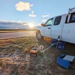 An image with clouds and the sun setting in the horizon. A white truck is parked on the right in a field setting. Next to the truck is sampling equipment next to a drain.