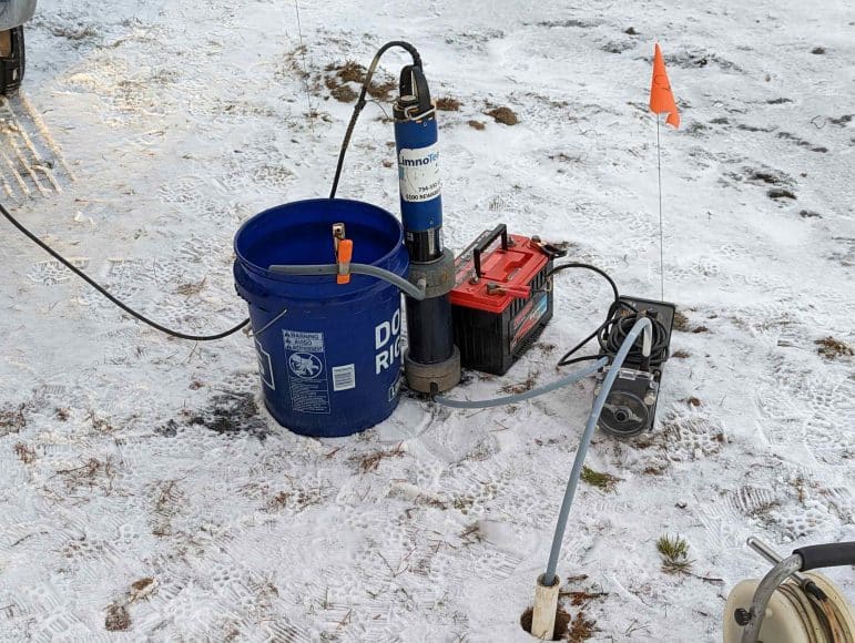 Image shows sampling equipment on a snowy ground. There is a bucket that has a clamp holding tubing connected to a sampler. The tubing is threaded through the equipment and is placed into the ground through a pvc pipe. There is a battery to provide power.