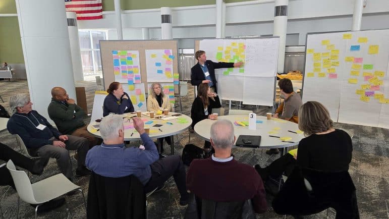 A group of people sitting at tables participating in the University of Michigan Urban Collaboratory Water and Energy Summit in 2025. A person is standing in front of white boards with sticky notes of different colors while the people at the table engage in the discussion.
