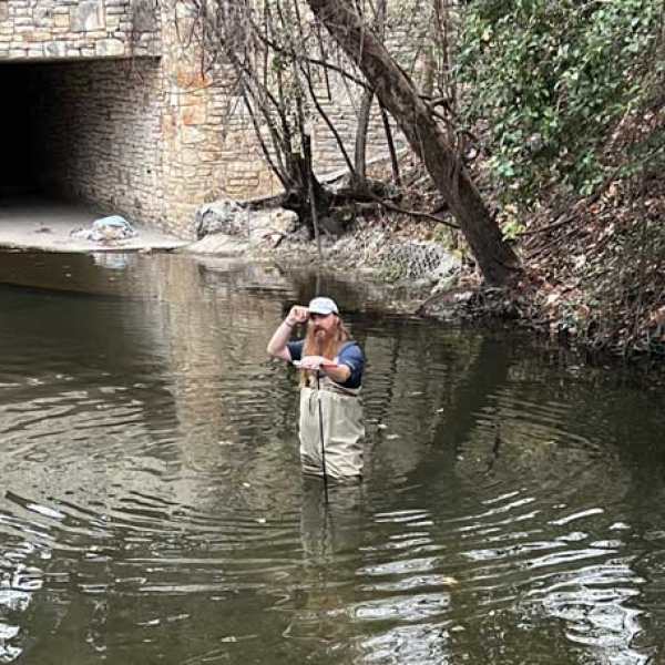 A man standing in water knee deep in Waller Creek taking measurements.